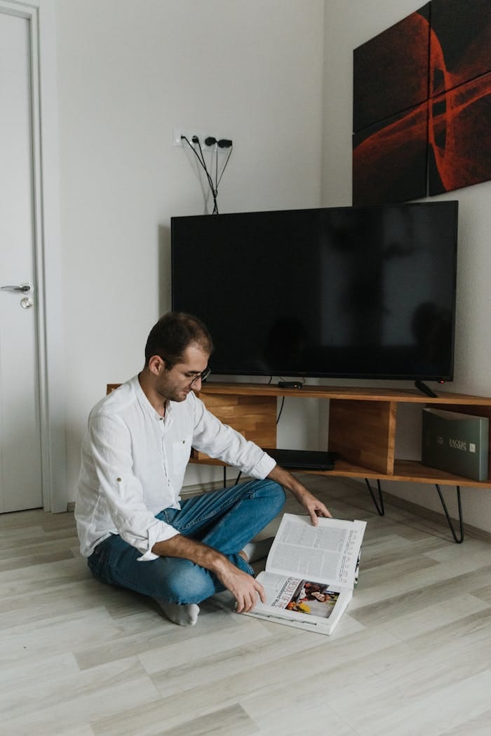 A man in a white shirt reads a magazine on the floor of a modern living room.